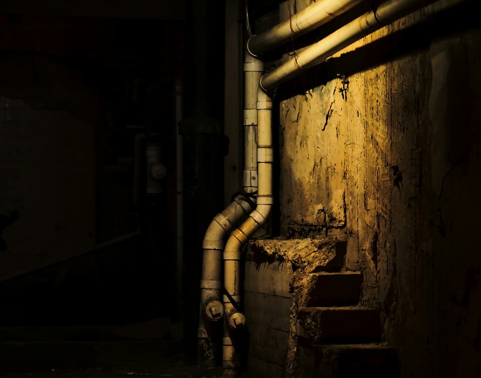 Pale pipes running down a stained basement wall beside concrete steps